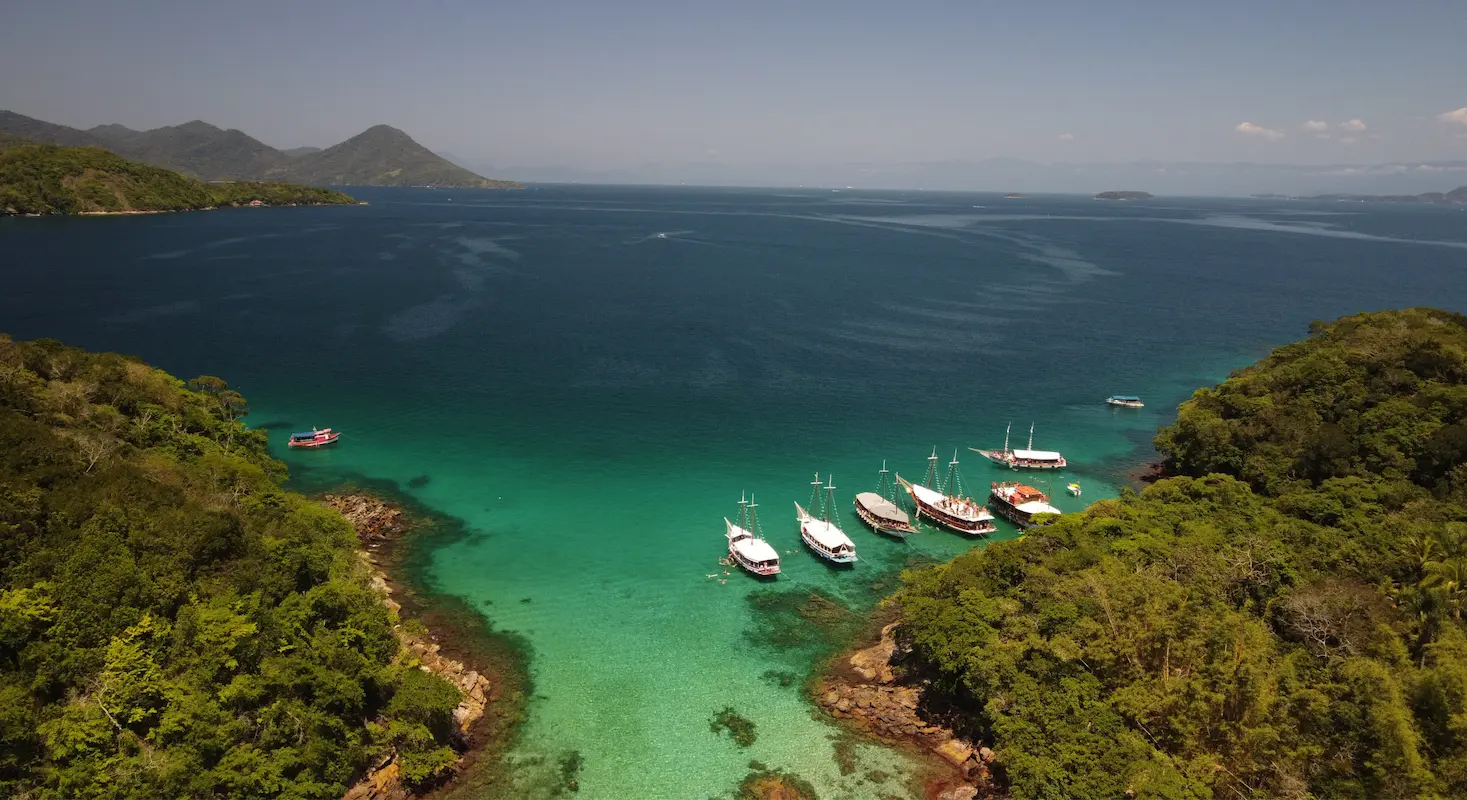 Vista aérea de Angra dos Reis com águas cristalinas, barcos ancorados, ilhas verdes e montanhas ao fundo.