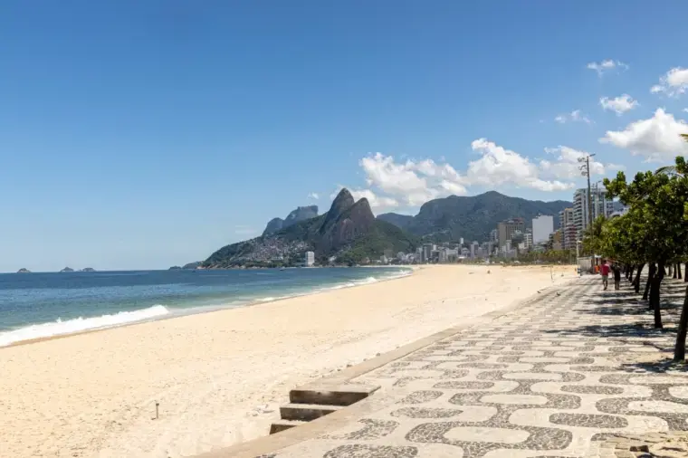 Vista da praia do Leblon com calçadão arborizado, areia ampla, mar azul e montanhas ao fundo.